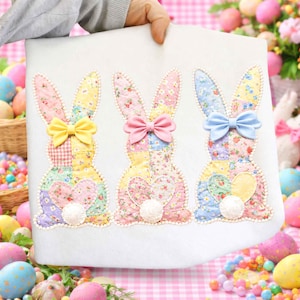 May include: A white fabric panel with three appliqued bunny rabbits, ideal for Easter. Each bunny has a patchwork body, a bow, and a fluffy tail. The bows are yellow, pink, and blue. The background is decorated with Easter eggs and flowers.