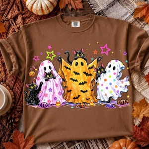 May include: A brown t-shirt featuring a graphic of three black cats wearing ghost costumes. One cat is in a pink floral sheet, one in a yellow bat-patterned sheet, and one in a white polka-dot sheet. Colorful stars and candy are scattered around.