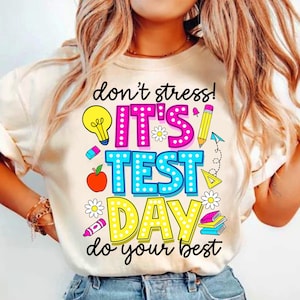 Pode incluir: Camiseta creme com um gráfico colorido que diz "don't stress! It's test day do your best." O design inclui uma lâmpada, um lápis, uma maçã e outros elementos escolares em cores vibrantes.
