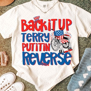 Könnte beinhalten: Cremefarbenes T-Shirt mit dem Text "Back it UP Terry Putin in Reverse" in Rot und Blau. Das Design beinhaltet einen Rollstuhl, der mit der amerikanischen Flagge und einem patriotischen Hut verziert ist.