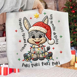 May include: A white Christmas-themed embroidered fabric featuring a cartoon donkey wearing a Santa hat and wrapped in colorful Christmas lights. Text includes phrases in Spanish. Red and green star and cross accents surround the donkey.