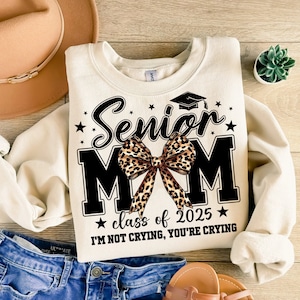 May include: A cream-colored sweatshirt with the text "Senior Mom" in black with a leopard print bow. The text "class of 2025" is below the bow and the text "I'm not crying, you're crying" is below that.