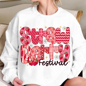 May include: White sweatshirt with a red, pink, and white graphic design that says "Strawberry Festival". The graphic is made up of different patterns and includes strawberries.
