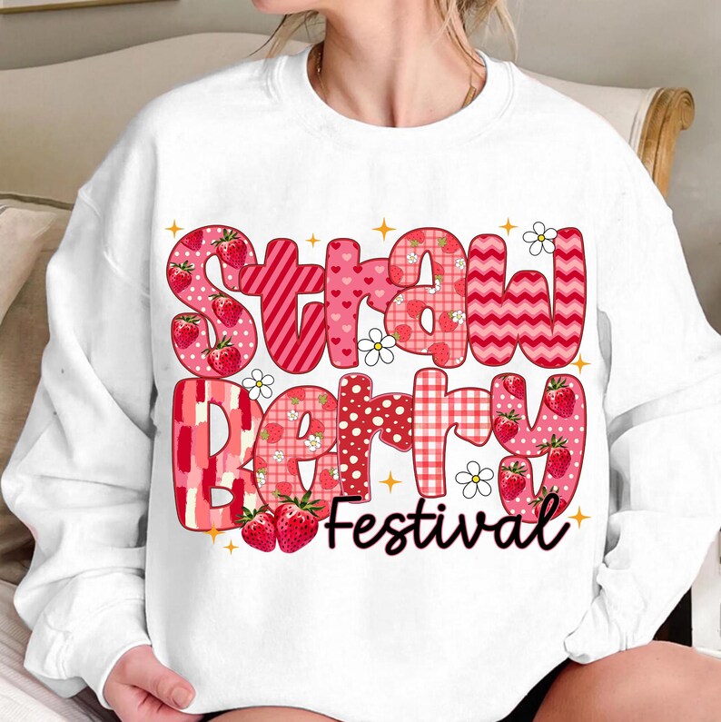 May include: White sweatshirt with a red, pink, and white graphic design that says "Strawberry Festival". The graphic is made up of different patterns and includes strawberries.