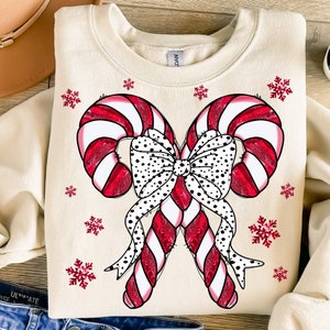 May include: A cream-colored sweatshirt with a red and white candy cane design with a black and white polka dot bow. The candy canes are crossed and tied together with the bow. There are small red snowflakes scattered around the design.