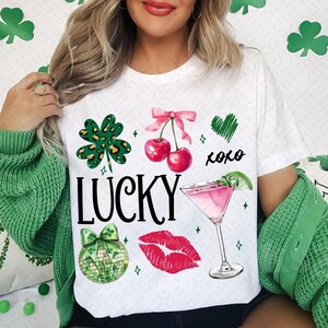 May include: White t-shirt with a green shamrock, pink cherries, a pink cocktail, a red lip, and the text "LUCKY xoxo".