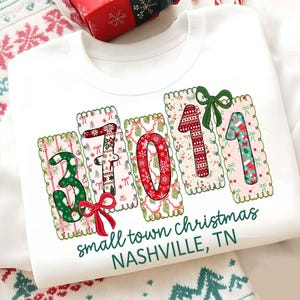 Puede incluir: Sudadera blanca con un diseño navideño. El diseño presenta los números "37011" en una fuente festiva, con elementos decorativos como copos de nieve y lazos. El texto "small town christmas NASHVILLE, TN" está impreso debajo.