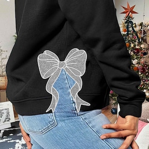 Puede incluir: Sudadera negra con un gran diseño de lazo plateado brillante en la espalda. El lazo tiene una apariencia texturizada. La sudadera se combina con unos vaqueros azules.