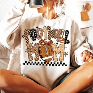 May include: A cream-colored sweatshirt with the words "FOOTBALL MOM" in a mix of patterns and colors. The word "FOOTBALL" has a checkered pattern, leopard print, and stars. A football with a bow is in the center. A black and white checkered border is at the bottom.