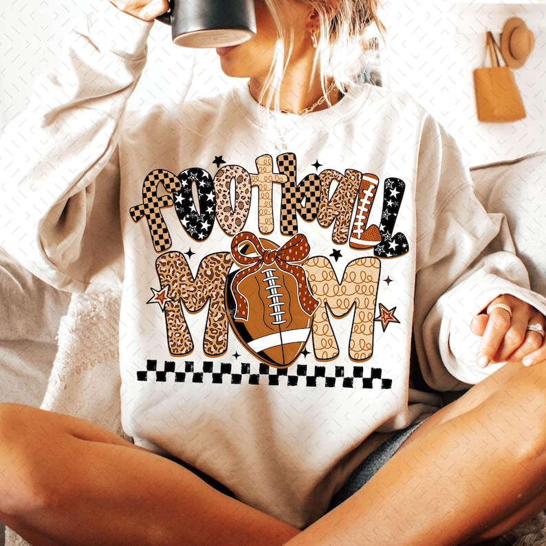 May include: A cream-colored sweatshirt with the words "FOOTBALL MOM" in a mix of patterns and colors. The word "FOOTBALL" has a checkered pattern, leopard print, and stars. A football with a bow is in the center. A black and white checkered border is at the bottom.