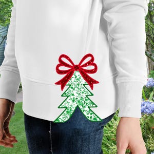 Może przedstawiać: Biała bluza z zielonym wzorem choinki z czerwoną kokardką na górze. Choinka jest ozdobiona białym wzorem kwiatowym.