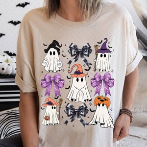 Puede incluir: Una camiseta beige con un dise&ntilde;o de Halloween con varios fantasmas que llevan sombreros y sostienen varios objetos. Los fantasmas est&aacute;n rodeados de lazos morados y negros con lunares blancos.