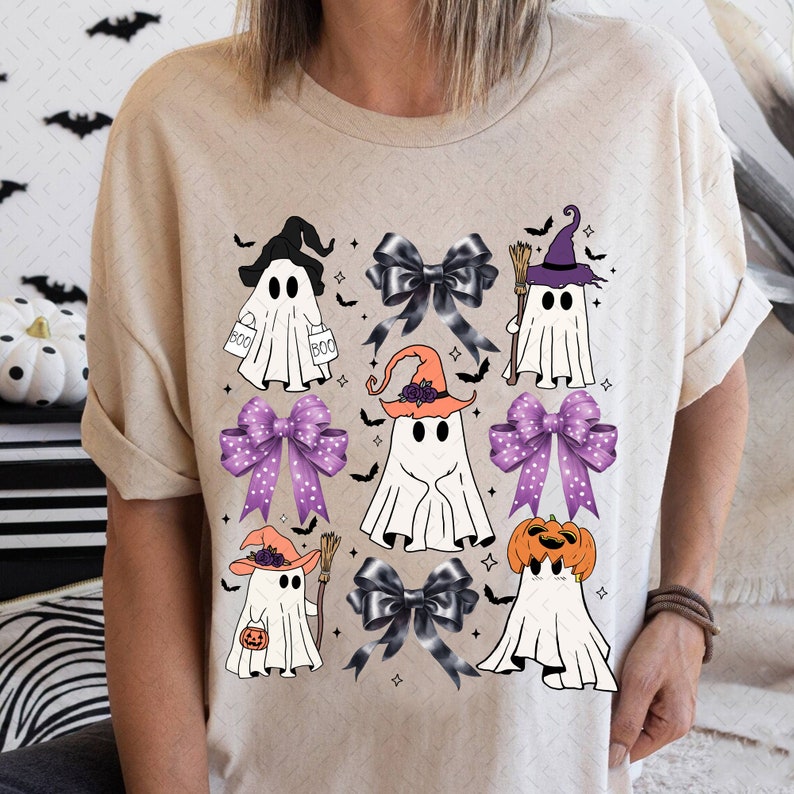 Puede incluir: Una camiseta beige con un dise&ntilde;o de Halloween con varios fantasmas que llevan sombreros y sostienen varios objetos. Los fantasmas est&aacute;n rodeados de lazos morados y negros con lunares blancos.