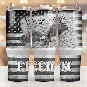 May include: A stainless steel tumbler with a white handle, featuring a black and white American flag design, the text "We the People", and a bald eagle illustration. The word "FREEDOM" is printed at the bottom.