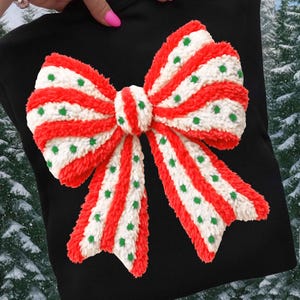May include: Black garment featuring a large, textured bow. The bow has red and white stripes with green polka dots. The bow is the focal point, with a wintery background visible.