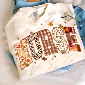 May include: Cream-colored sweatshirt with the word "NURSE" in a patchwork design. The letters feature patterns like leopard print, plaid, and floral prints, with autumn leaves and stars. The shirt is folded on top of blue jeans.