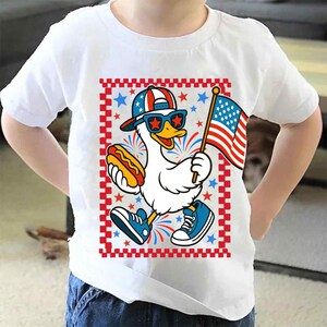 May include: White t-shirt with a cartoon duck wearing sunglasses and a red, white, and blue hat holding a hot dog and an American flag. The duck is standing on a checkered background with fireworks.