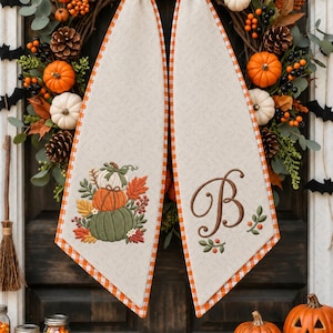 May include: Two beige fabric door hangers with orange and white checkered trim. The left hanger features an embroidered stack of pumpkins with autumn leaves. The right hanger displays a large brown embroidered letter B with small leaf accents.