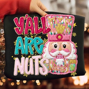 May include: Black tote bag with the text "Y'ALL ARE NUTS" in pink and turquoise. A colorful nutcracker design is featured on the right side. The nutcracker is pink, gold, and green, with a white beard and a crown.