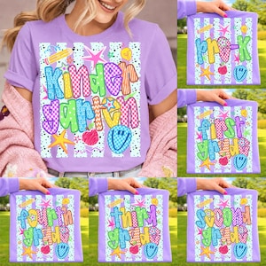 May include: A light purple t-shirt with a colorful "Kindergarten" graphic. The design includes stars, pencils, and a smiley face. Additional shirts display "Pre-K", "First Grade", "Second Grade", "Third Grade", and "Fourth Grade" graphics.