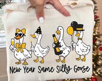 May include: Off-white sweatshirt featuring a whimsical design of four cartoon geese celebrating. Each goose is adorned with festive accessories like sunglasses, party hats, and champagne. The text "New Year Same Silly Goose" is printed below the geese.