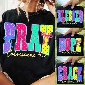 May include: Black t-shirts with colorful text designs. One shirt says "PRAY" with "Colossians 4:2". Other shirts say "BLESSED" with "John 20:29", "HOPE" with "Psalm 130:5", and "GRACE" with "2 Corinthians 12:9".