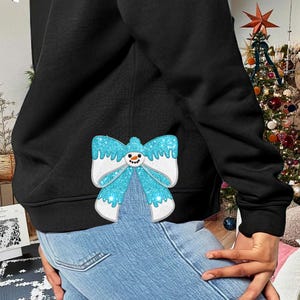 May include: A black hoodie with a blue and white glitter snowman bow on the front. The hoodie is worn by a person with blue jeans.