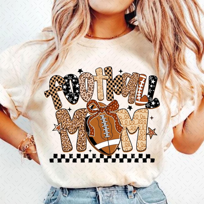 May include: Cream-colored t-shirt with the words "FOOTBALL MOM" in a mix of fonts and patterns. The design includes a football with a bow, stars, and a checkered border. The text is in black, brown, and white.