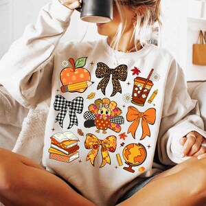 May include: Cream-colored sweatshirt featuring a collection of fall-themed graphics. The design includes an apple with a pencil, bows, a turkey, a coffee cup, books, a globe, and fall leaves. The graphics are in shades of orange, brown, black, and white.