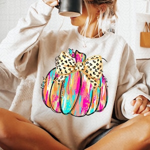 May include: A white sweatshirt with a colorful abstract pumpkin design and a leopard print bow. The pumpkin is painted with pink, blue, yellow, and orange colors.