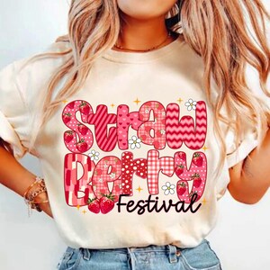 May include: White t-shirt with a pink and red graphic that says "Strawberry Festival". The graphic is made up of different patterns and includes strawberries.