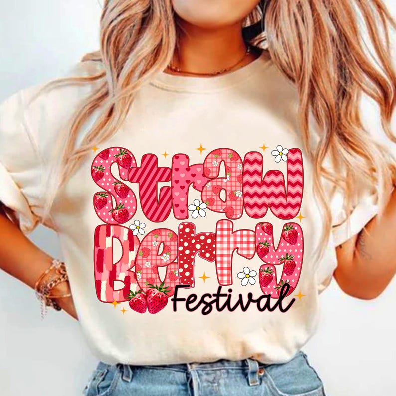 May include: White t-shirt with a pink and red graphic that says "Strawberry Festival". The graphic is made up of different patterns and includes strawberries.