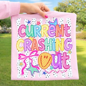 May include: A light pink t-shirt featuring a graphic print with the text "CURRENT CRASHING OUT" in colorful, checkered, and patterned letters. The design includes a yellow smiley face, pink bows, and various stars against a black polka dot background.