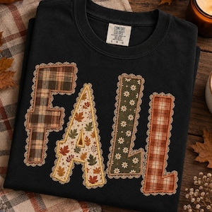 May include: A black crewneck t-shirt featuring the word FALL in large, appliqued letters. Each letter has a unique pattern: brown plaid, autumn leaves on cream, white daisies on dark green, and orange plaid, all bordered with tan lace trim.