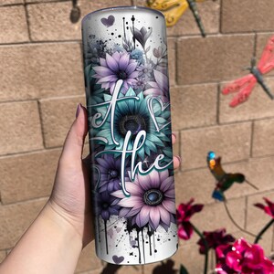 May include: A white tumbler with a floral design and the text "Let the" in black. The flowers are purple, blue, and green. The tumbler has a black and white splatter paint design.