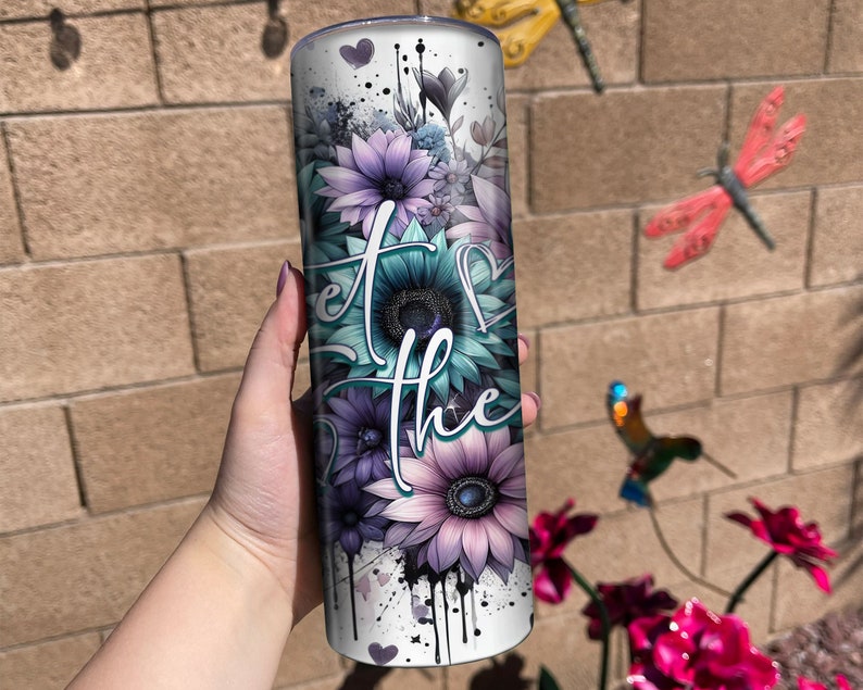May include: A white tumbler with a floral design and the text "Let the" in black. The flowers are purple, blue, and green. The tumbler has a black and white splatter paint design.