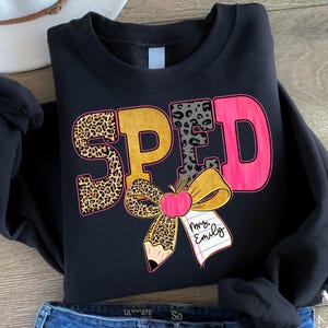 May include: Black sweatshirt featuring the word "SPED" in leopard print, gold, grey, and pink. A bow with a pencil and apple design is below the letters, with the name "Mrs. Emily" on a notepad. The sweatshirt is folded.