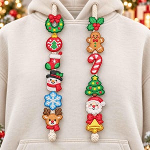 Può includere: Una felpa con cappuccio beige dotata di due spessi cordini decorati con toppe ricamate a tema natalizio. Il cordino sinistro mostra una ghirlanda, una pallina, una calza, un pupazzo di neve, un fiocco di neve e una renna. Il destro mostra agrifoglio, omino di pan di zenzero, bastoncino di zucchero, albero, Babbo Natale e una campana.