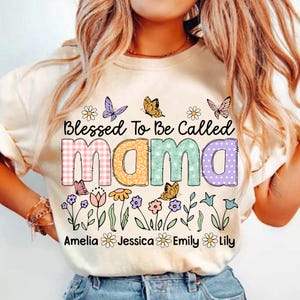 Puede incluir: Una camiseta blanca con el texto "Blessed To Be Called Mama" en letras de colores con un dise&ntilde;o de patchwork. Las letras est&aacute;n rodeadas de flores y mariposas. Los nombres Amelia, Jessica, Emily y Lily est&aacute;n impresos debajo del texto.