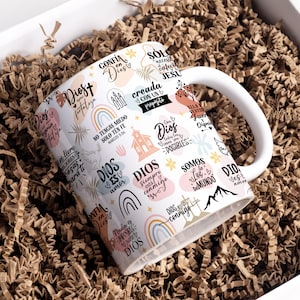 May include: White ceramic mug with a colorful design featuring religious quotes and phrases in Spanish. The mug has a handle and is filled with brown packing peanuts.