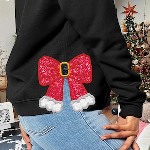 May include: Black hoodie with a red glitter bow with a gold buckle on the back. The bow has white fluffy trim around the edges.