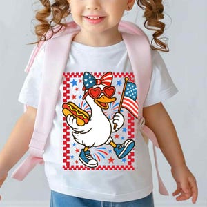 May include: White t-shirt with a cartoon image of a duck wearing red heart-shaped sunglasses and a red, white, and blue bow. The duck is holding a hot dog in one hand and an American flag in the other. The background is a red, white, and blue checkered pattern with stars.