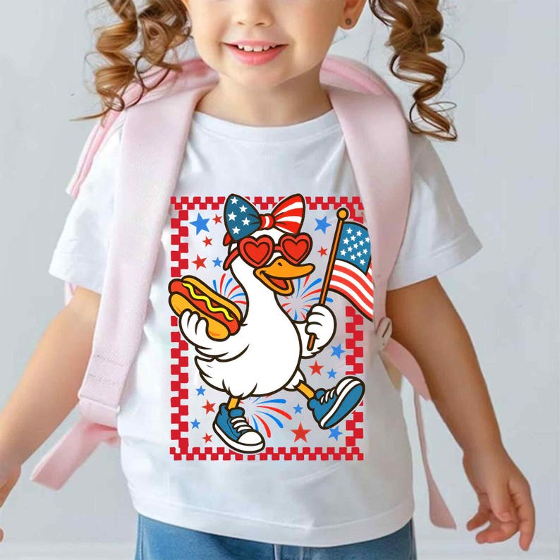 May include: White t-shirt with a cartoon image of a duck wearing red heart-shaped sunglasses and a red, white, and blue bow. The duck is holding a hot dog in one hand and an American flag in the other. The background is a red, white, and blue checkered pattern with stars.