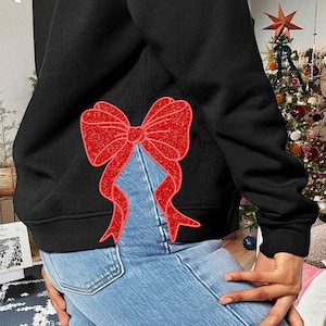 May include: A black hoodie with a red glitter bow on the back. The hoodie is worn over blue jeans.