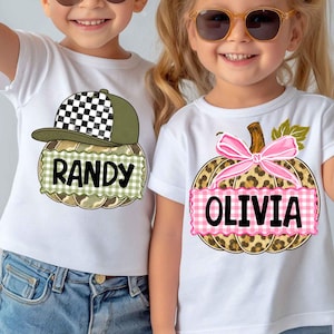 Op de afbeelding: Twee witte t-shirts met pompoenafbeeldingen. Het linker t-shirt toont een pompoen met een geblokte baseballpet en de naam RANDY. Het rechter t-shirt toont een pompoen met een roze strik en de naam OLIVIA, beide met bedrukte banners.