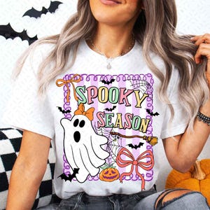 Op de afbeelding: Wit T-shirt met een kleurrijke "Spooky Season" afbeelding. Het ontwerp bevat een spook met een strik, vleermuizen, een pompoen en een heksenbezem. De afbeelding is omlijst met een paars-witte gestreepte rand.