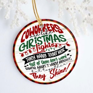 May include: Round Christmas ornament with a white woodgrain background and red and black buffalo plaid trim. The ornament features the text "Coworkers are like Christmas lights. They hang together. Half of them don't work, some aren't very bright, but together they shine!"