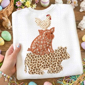May include: White sweatshirt featuring appliqued farm animals: a chicken, pig, and cow. The chicken has a polka dot pattern, the pig has a floral pattern, and the cow has a leopard print. The sweatshirt is held by a person.