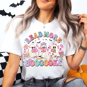 May include: White t-shirt with a colorful graphic featuring three ghost characters reading books. The text "READ MORE BOOOOOKS" is arched above the ghosts. The design includes flowers, bats, and spiderwebs.
