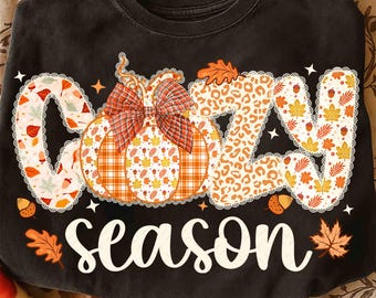 May include: Black sweatshirt featuring the words "COZY SEASON" in a seasonal design. The letters are decorated with autumn leaves, acorns, and a pumpkin with a plaid bow. A comfortable, casual style.